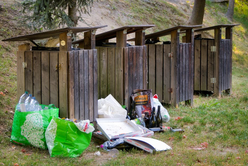 A row of four outdoor wooden rubbish bins with sloped metal lids situated on a grassy area near a sloped hillside with trees and bushes in the background. The bins are dark brown with weathered, rough