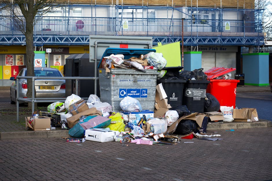 A pile of discarded rubbish, including black plastic trash bags, cardboard, and miscellaneous debris, is stacked against a weathered red metal door with graffiti and security signs. The door, set into