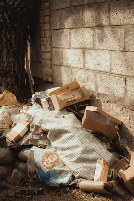 Two large, crumpled black plastic rubbish bags, slightly shiny and weathered, sit on the edge of a paved sidewalk in front of a dark metal fence with vertical bars. The bags are filled with waste mate
