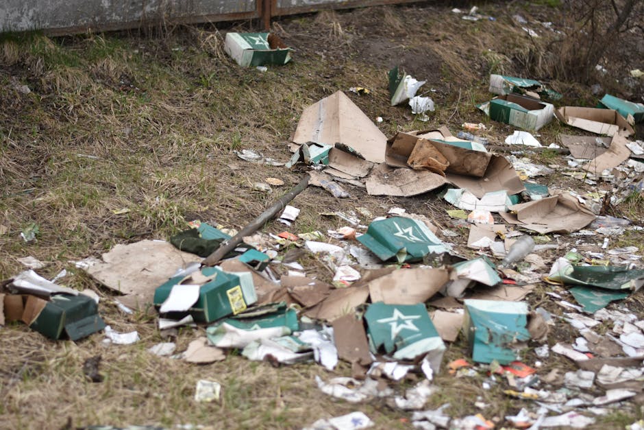 An aerial view of a large outdoor area strewn with various types of rubbish, including plastic bags, cardboard boxes, and discarded household items. Several workers are actively sorting through the de
